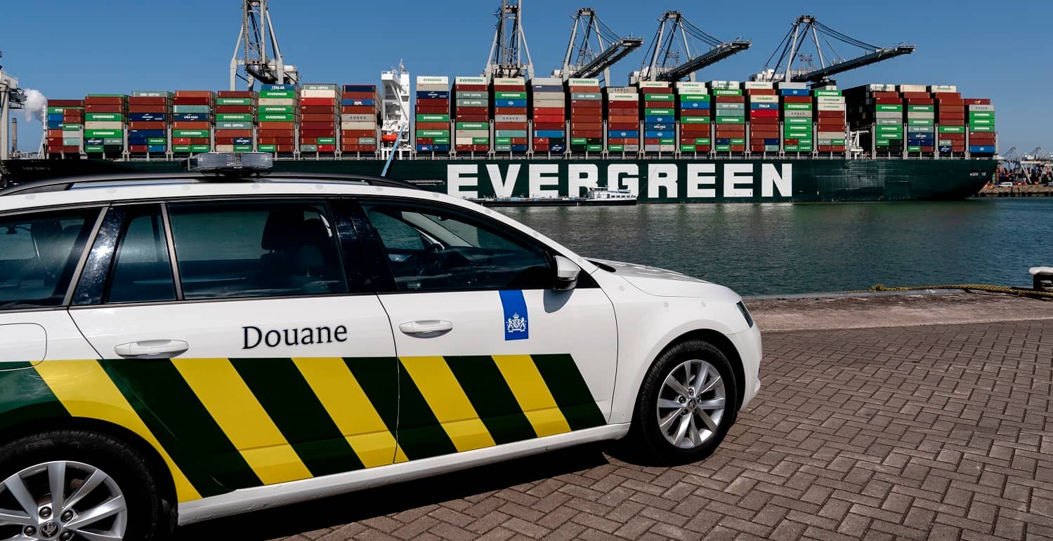 Een auto van de Douane in de haven, met op de achtergrond een containerschip. 