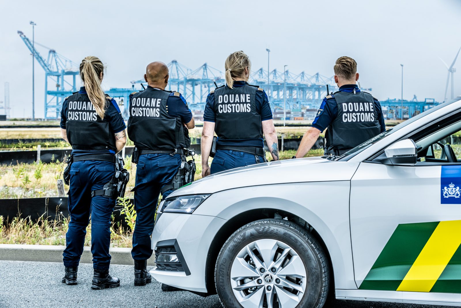 Vier medewerkers van de Douane in uniform, met hun rug richting de camera en een auto van de Douane op de voorgrond. 