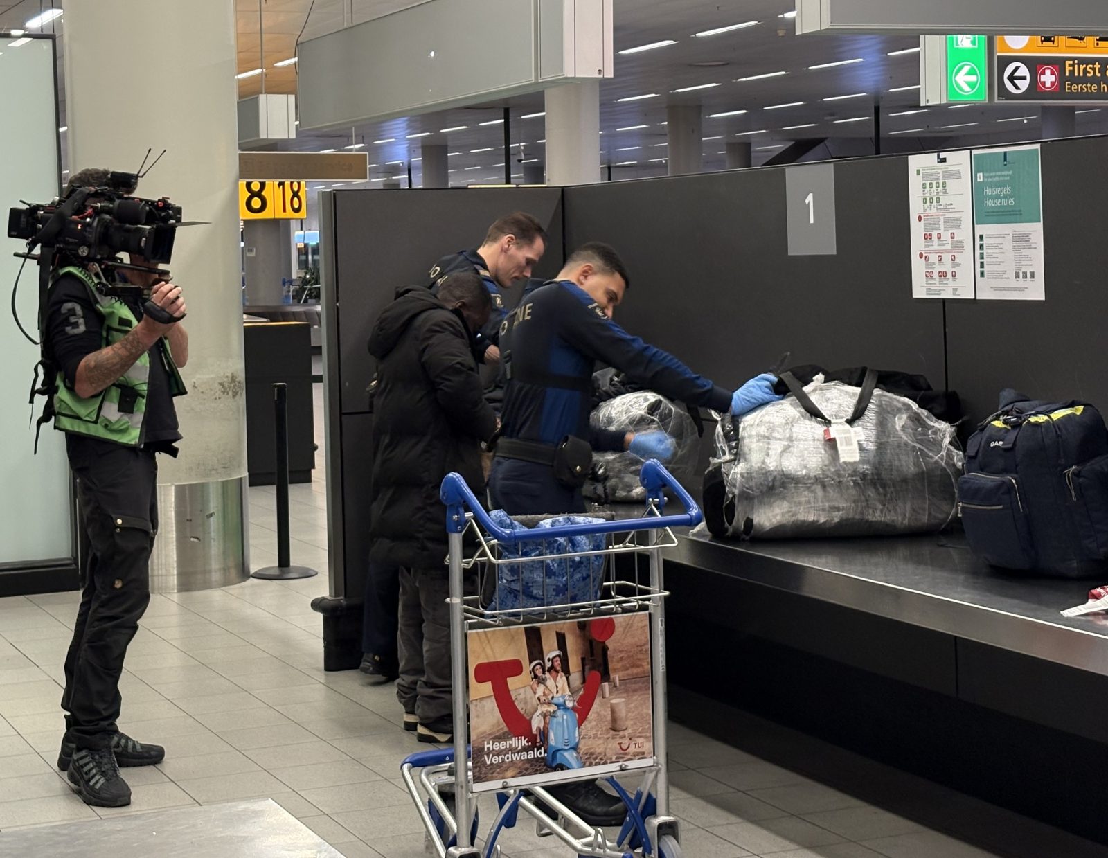 Twee douaniers controleren bagage op Schiphol terwijl ze door een cameraman gefilmd worden. 