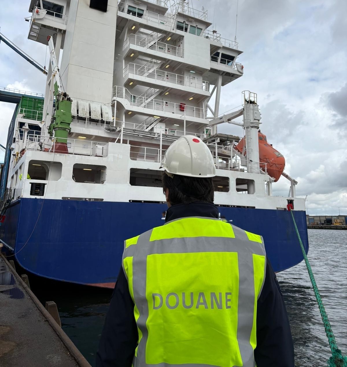 Douanemedewerker met veiligheidshelm op en geel hesje aan, loopt richting containerschip.