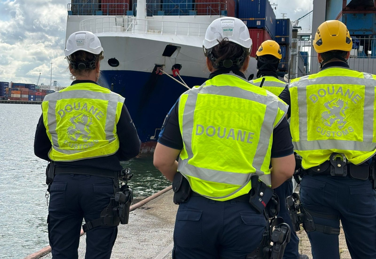 Vier douanemedewerkers met veiligheidshelmen op en gele hesjes aan staan voor een containerschip.